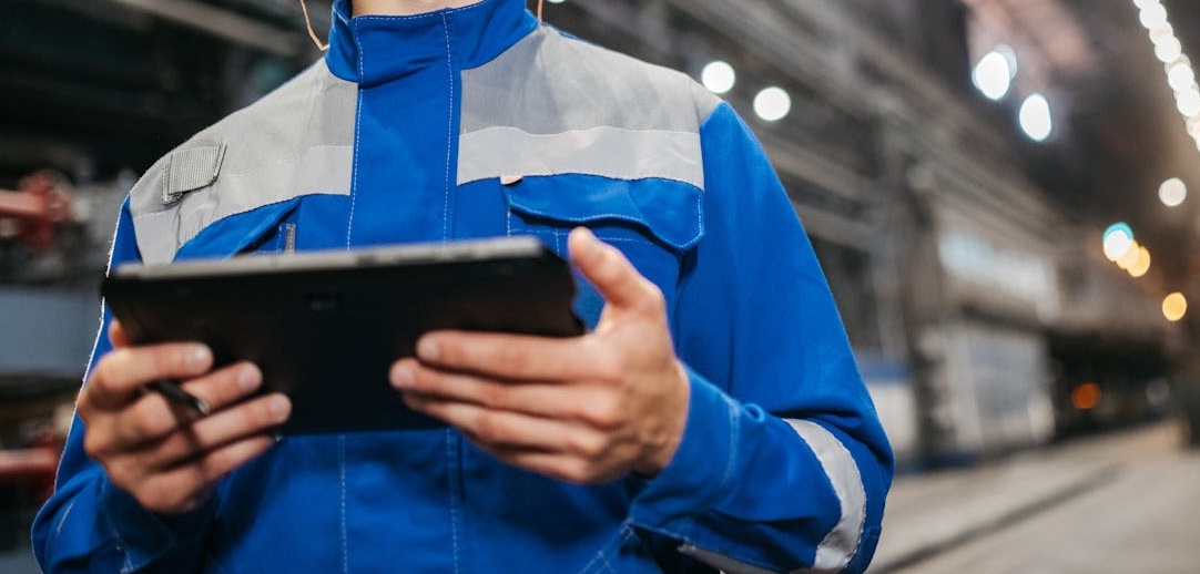 A man in a warehouse wearing a blue and grey jacket and holding a tablet. 